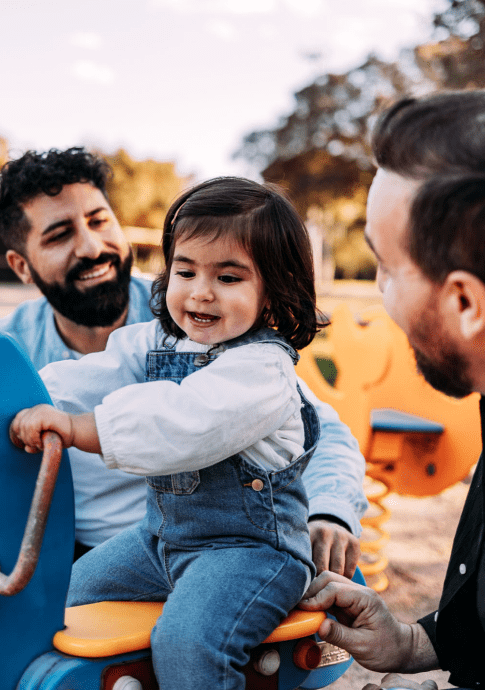 Male couple playing with their daughter on a seesaw in the park.