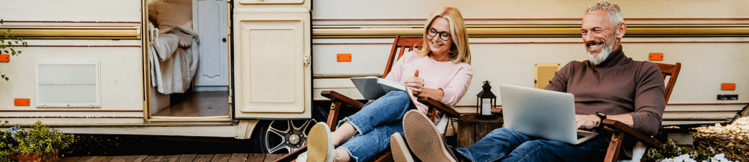 cheerful senior couple talking in the camper van yard sitting on deck chairs with book and laptop