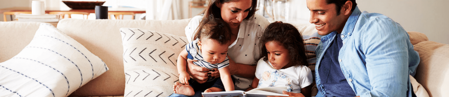 Hispanic couple sitting on the sofa reading a book at home with their baby son and young daughter